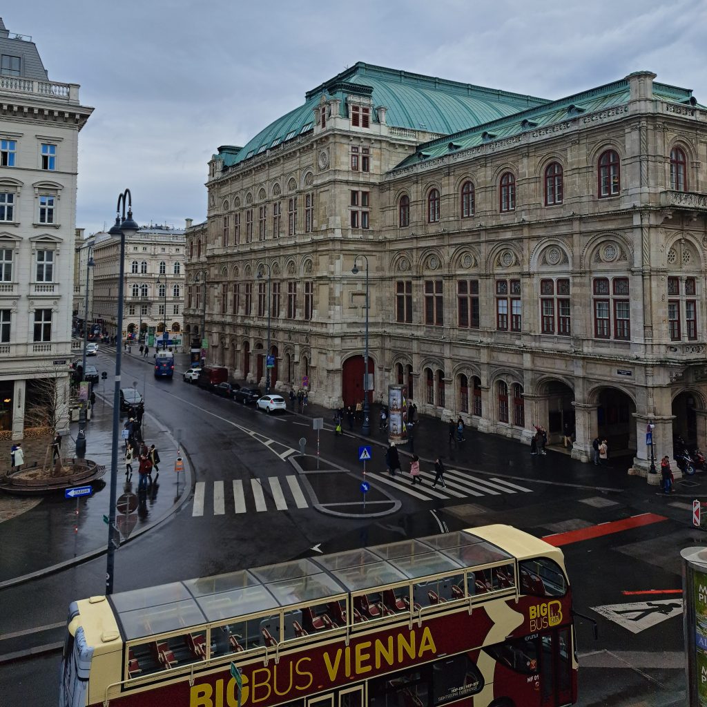Görselde Wiyana sokakları, tarihi binalar ve üstü açık bir tur otobüsü yer almakta. Hava kapalı ve ıslak bir zemin gözlemleniyor.