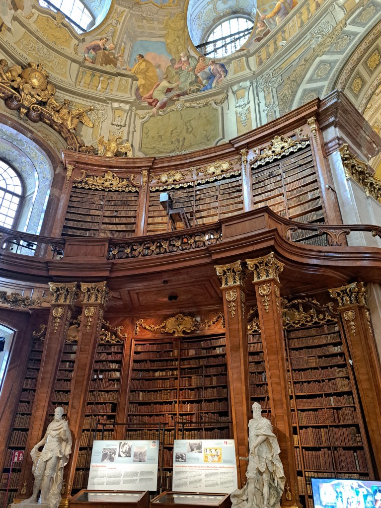 Viyana'daki tarihi kütüphanenin iç mekan görüntüsü, ahşap raflarda kitaplar ve tablolarla süslenmiş tavan.